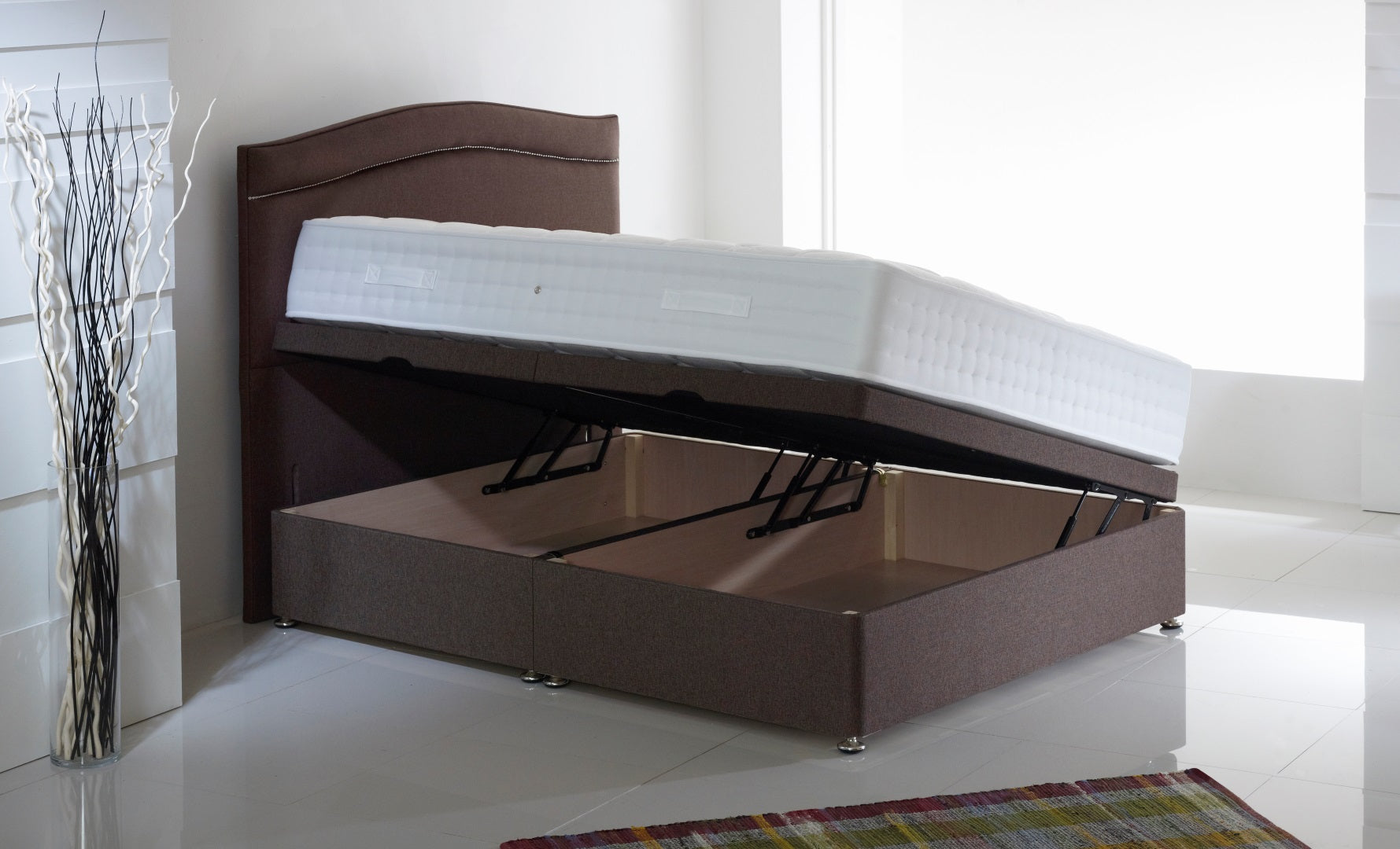 Picture of a side lift single ottoman in a white room with a brown headboard, a rug on the floor and a vase filled with white and black sticks to the side of it