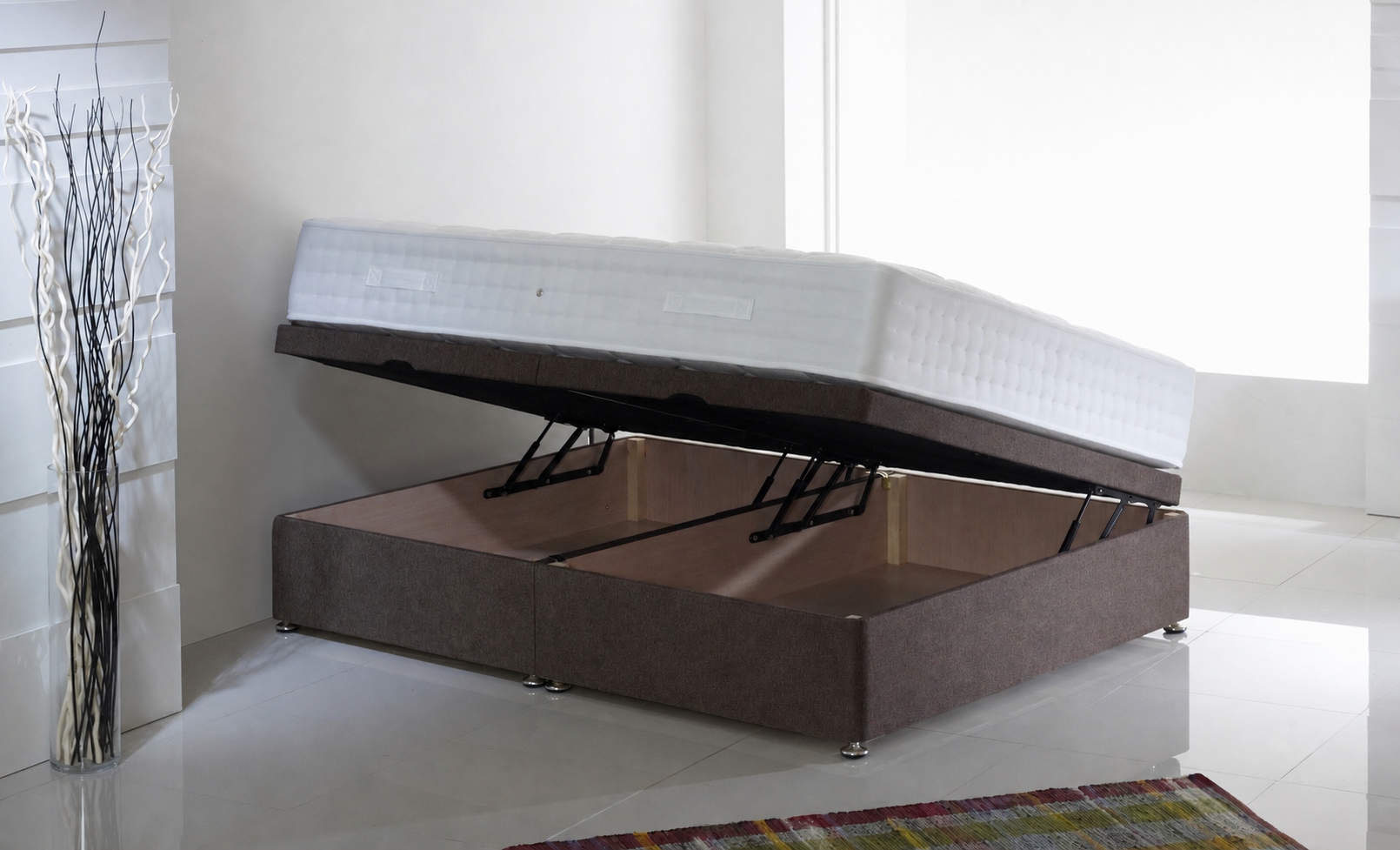 Picture of a side lift single ottoman in a white room with a brown headboard, a rug on the floor and a vase filled with white and black sticks to the side of it